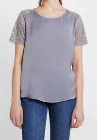 Blusa gris de manga corta con detalles de encaje, tejido suave, escote redondo y corte holgado. Combinada con jeans azul oscuro.