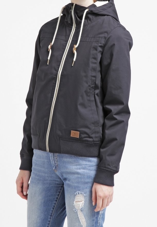 Svart jacka med huva, vita dragsko, zip fram, ribbade ärmar och en brun logotyp-patch. Bärs med ljusblå trasiga jeans.