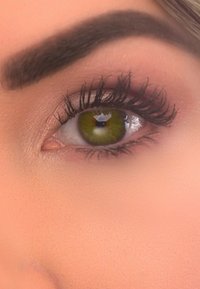 Nahaufnahme eines Auges mit grünem Iris und langen schwarzen Wimpern, detaillierte Hautstruktur um das Auge, mit einem hellrosa Hintergrund.