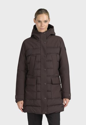 Femme portant un manteau d'hiver matelassé brun foncé avec une capuche, des boutons-pression sur le devant et plusieurs grandes poches, debout devant un fond uni.