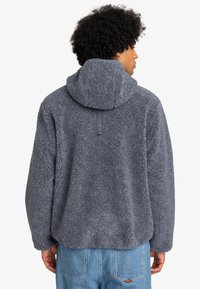Pull en polaire gris avec capuche, présentant un extérieur texturé et duveteux. Design simple, avec une vue partielle de l'arrière et un détail de cordon de serrage.