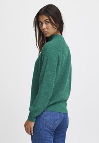 Groene, gestructureerde gebreide trui met een hoge hals en lange mouwen. De achterkant toont een relaxte pasvorm, gecombineerd met blauwe spijkerbroek.