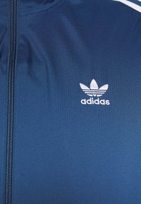 Blue Adidas jacket with white embroidered Trefoil logo and zipper running vertically on the left side.
