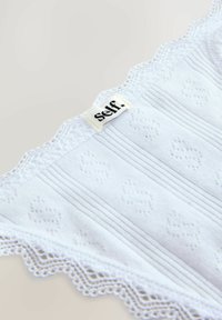 White cotton garment with ribbed texture and scalloped lace trim. Features the label “self.” stitched at the top.