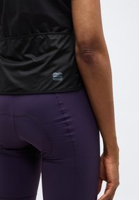 Κοντινή λήψη της κάτω πλάτης και του δεξιού βραχίονα ενός ατόμου που φοράει μαύρο τοπ από δίχτυ και μοβ ποδηλατικά σορτς με ραμμένο επένδυση.