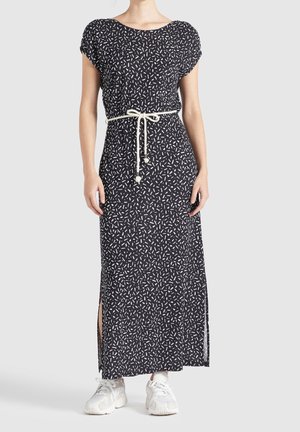 Femme portant une robe longue noire avec un imprimé abstrait blanc, ceinture en corde, fentes latérales et baskets blanches, debout devant un fond uni.