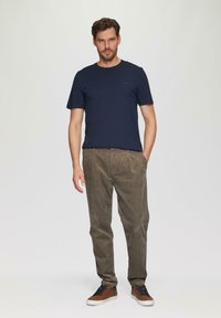 Mann steht mit den Händen in den Taschen, trägt ein dunkelblaues T-Shirt, braune Cordhosen und braune Sneaker mit weißen Sohlen.