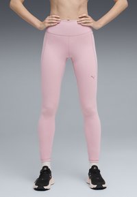 Egy magas derekú rózsaszín leggings-et viselő és fekete futócipőt hordó személy áll, kezével a csípőjén, szürke háttér előtt.
