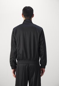 Black jacket with a stand collar, elasticated cuffs, and waistband. Smooth texture with a clean, minimalistic design. Back view.