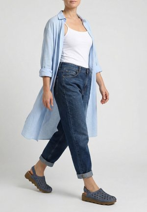 Kobieta idąca, ubrana w podwinięte ciemnoniebieskie dżinsy, biały top, jasno niebieską długą koszulę i niebieskie, perforowane sandały typu slingback z brązowymi podeszwami.