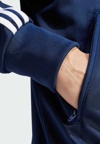 Veste de sport marine avec des rayures blanches sur les manches, en tissu texturé, et une poche zippée sur le côté. Gros plan sur la manche et la main dans la poche.