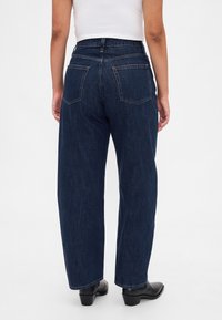 Σκούρο μπλε τζιν με ψηλή μέση, χαλαρή εφαρμογή, δύο πίσω τσέπες και εμφανείς ραφές, σε συνδυασμό με μαύρα μποτάκια στο αστράγαλο.