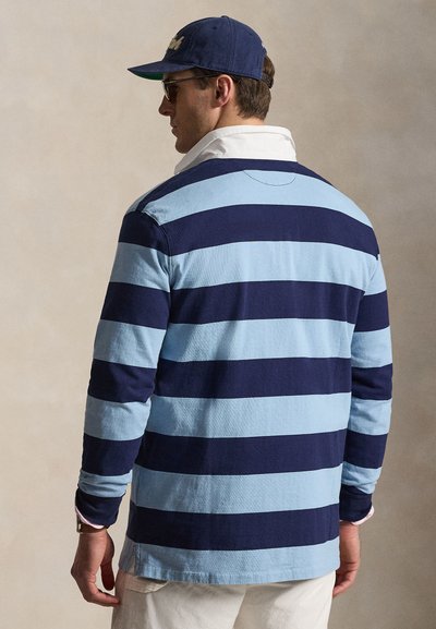 Homme portant un maillot de rugby à manches longues rayé bleu marine et bleu clair avec col blanc et casquette de baseball bleu marine, vu de dos.