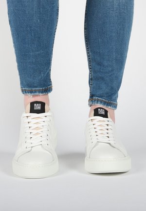 Hvide snøresneakers med mærket "BLACK STONE" båret med opbukket blå jeans, vist forfra på en ensfarvet lys baggrund.