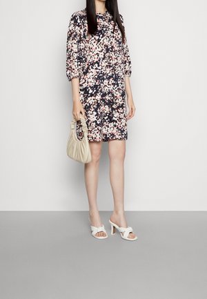 Frau trägt ein knielanges Blumenkleid, hält eine beige geraffte Handtasche und weiße Sandaletten mit Absatz vor einem einfarbigen Hintergrund.