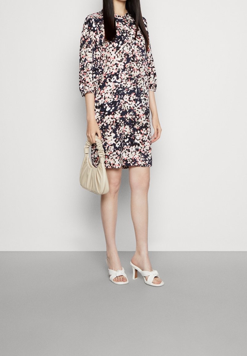 Femme portant une robe fleurie longueur genou, tenant un sac à main beige froncé et des sandales blanches à talons, sur un fond uni.