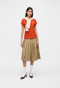 Κόκκινο εφαρμοστό T-shirt με λογότυπο, κρεμ κασκόλ με υφή δεμένο στον λαιμό, φουστάνι midi με πιέτες σε ταμπά απόχρωση, λευκές κάλτσες και μπορντό παπούτσια με τακούνι.