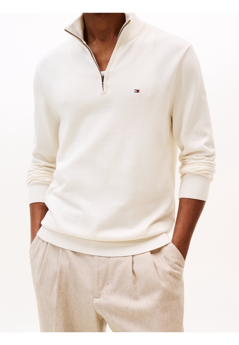 Homme portant un pull crème à glissière quart de fermeture avec un petit logo et un pantalon plissé beige, mains dans les poches, sur fond blanc.