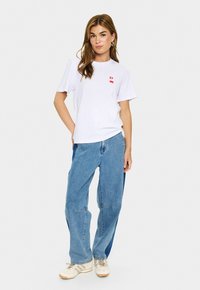 Weißes Baumwoll-T-Shirt mit einem kleinen roten Grafik, kombiniert mit lockeren blauen Denim-Hosen mit entspannter Passform und minimalen Hardware-Details.