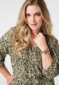 Chemise à motifs vert et crème avec un devant à boutons, des manches longues et une ceinture à la taille. Présente des bracelets dorés et un accent de bijou délicat.