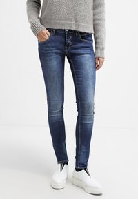 Person som bär blå skinny jeans, en grå stickad tröja och vita platåskor, står med en hand i fickan.