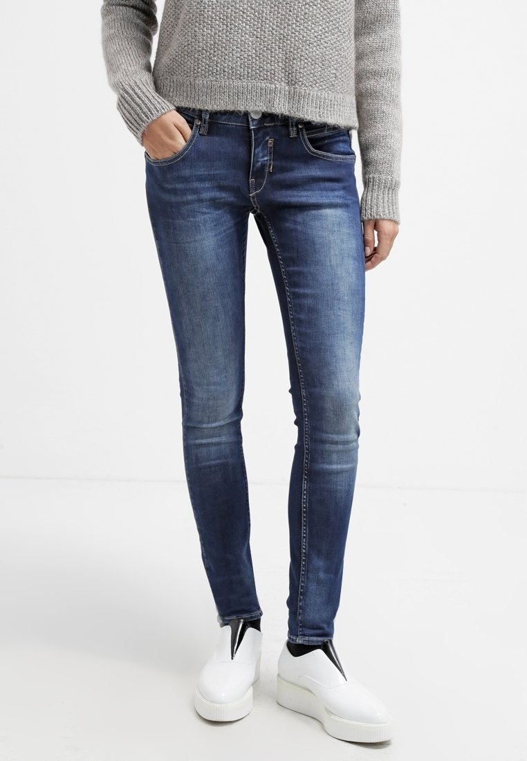 Person som bär blå skinny jeans, en grå stickad tröja och vita platåskor, står med en hand i fickan.