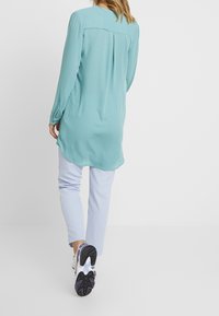 Chemise manches longues vert-bleu clair avec un pli au dos, associée à un pantalon bleu clair et des chaussures de sport blanches avec semelles noires et violettes.