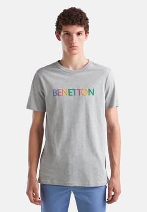 Joven con cabello rizado que lleva una camiseta gris clara con el texto colorido "BENETTON" y pantalones azules, de pie frente a un fondo blanco liso.