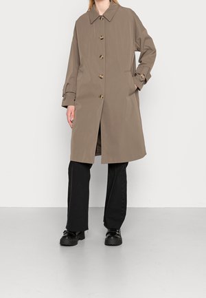 Persona con un abrigo largo beige de gabardina con detalles de botones, pantalones negros y zapatos negros gruesos, de pie frente a un fondo liso.