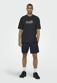 T-shirt noire avec impression graphique colorée, associée à un short bleu marine. Le mannequin porte des baskets noires et des chaussettes blanches. Fond uni.