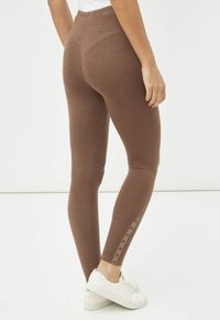 Leggings marrones de cintura alta hechos de una tela texturizada, con un detalle de botón en el costado. Combinados con zapatillas blancas.
