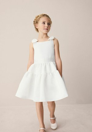 Junges Mädchen mit geflochtenen Haaren, das ein weißes ärmelloses Kleid mit Schleifen an den Schultern und weiße Mary-Jane-Schuhe trägt, stehend vor einem neutralen Hintergrund.