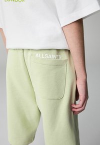 Svetlozeleni športni kratki hlače z strukturo, z zadnjim žepom in belim logotipom "ALLSAINTS". Nosi se z belo preveliko majico.