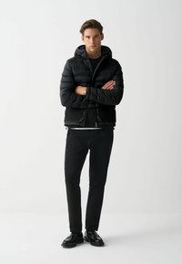 Schwarze Pufferjacke mit Kapuze, ausgestattet mit einem gesteppten Design und Reißverschluss. Kombiniert mit eng anliegenden schwarzen Hosen und schwarzen Schuhen.