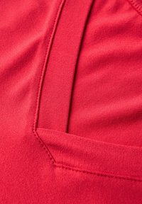 Czerwona tkanina o gładkiej teksturze, z detalem szwu podkreślającym kształt i design strefy kieszeni.