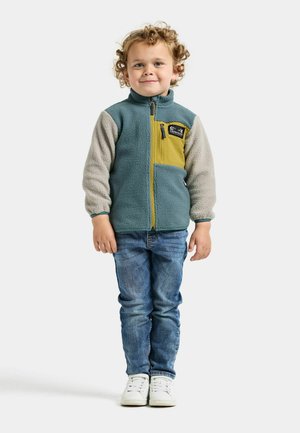 Fleecejack met een teal gekleurd lichaam, grijze mouwen en een gele borstzak. Ritssluiting en een logopatch aan de linkerkant. Kind draagt een spijkerbroek en witte sneakers.