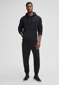 Felpa nera con cappuccio e coulisse, con tasca frontale, abbinata a pantaloni jogger neri. Il modello indossa sneakers nere con suola bianca.