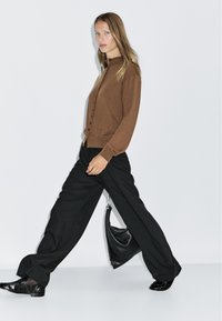Cardigan marrone con bottoni e maniche lunghe, abbinato a pantaloni neri a gamba larga, scarpe nere e una borsa nera. Design minimalista.