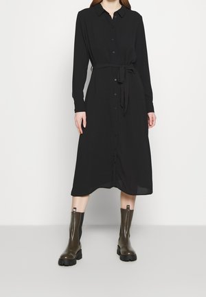 Personne portant une robe chemise midi noire à manches longues avec une ceinture nouée à la taille et des bottines épaisses vert foncé sur un fond clair.