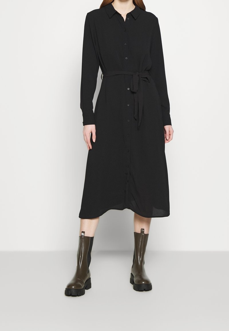 Personne portant une robe chemise midi noire à manches longues avec une ceinture nouée à la taille et des bottines épaisses vert foncé sur un fond clair.