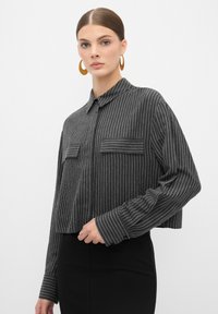 Geknipt, button-up shirt in donkergrijze stof met witte verticale strepen. Beschikt over twee voorzakken en knoopmanchetten.