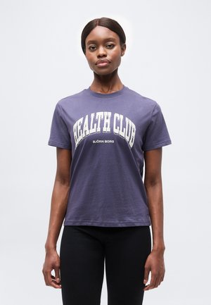 Frau mit dunklen Haaren in einem violetten "Health Club Björn Borg"-T-Shirt und schwarzen Hosen steht vor einem schlichten hellen Hintergrund.