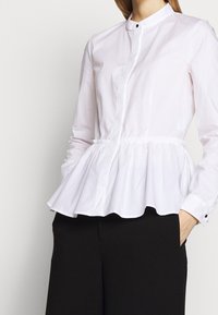 Donna che indossa una blusa peplum a maniche lunghe bianca con colletto alla cinese e pantaloni neri, con una mano in tasca.