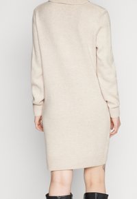 Abito maglione lavorato a maglia in beige chiaro, con maniche lunghe, colletto e polsini a costine, e orlo dritto. Texture morbida, design minimale.
