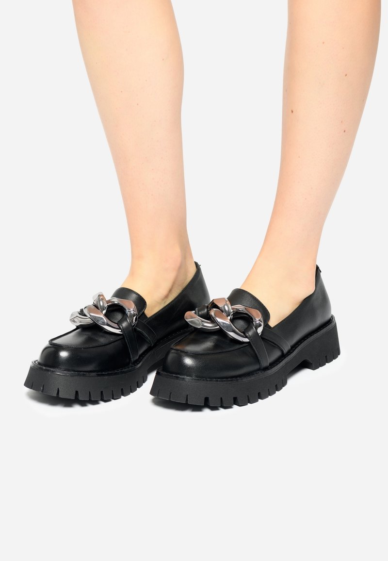 Mocassins à plateforme noire épaisse avec un détail de chaîne argentée surdimensionnée, portés pieds nus sur un fond clair.
