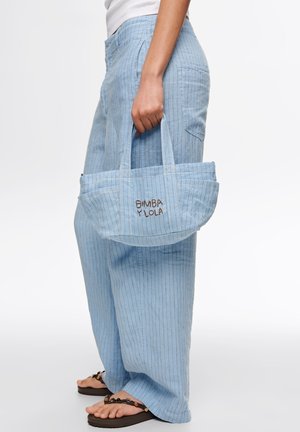 Persona con pantalones azul claro a rayas finas y sandalias, sosteniendo un pequeño bolso azul a rayas a juego con el texto "BIMBA Y LOLA".