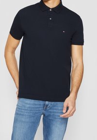 Man som bär en figursydd marinblå polo med knappar och ljusblå jeans, stående med en hand nära fickan.