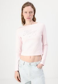 Lys rosa langermet crop top med rund hals og "Hugo" i rhinestones foran, brukt med lyseblå denim jeans.