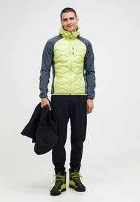 Puchowa kurtka w zielono-szarym materiale, z wysokim kołnierzem i zamkiem. Czarne spodnie oraz górskie buty z żółtymi akcentami. Trzyma czarną kurtkę.