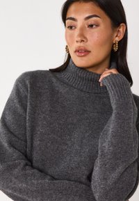 Maglione grigio lavorato a maglia con un collo a coste alto e maniche lunghe, che mostra una texture morbida e una vestibilità rilassata. Orecchini d'oro aggiungono un dettaglio contrastante.